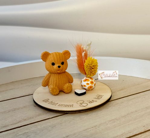 Ein brauner Bärenanhänger sitzt auf einem Holzbrett mit bunten Trockenblumen.